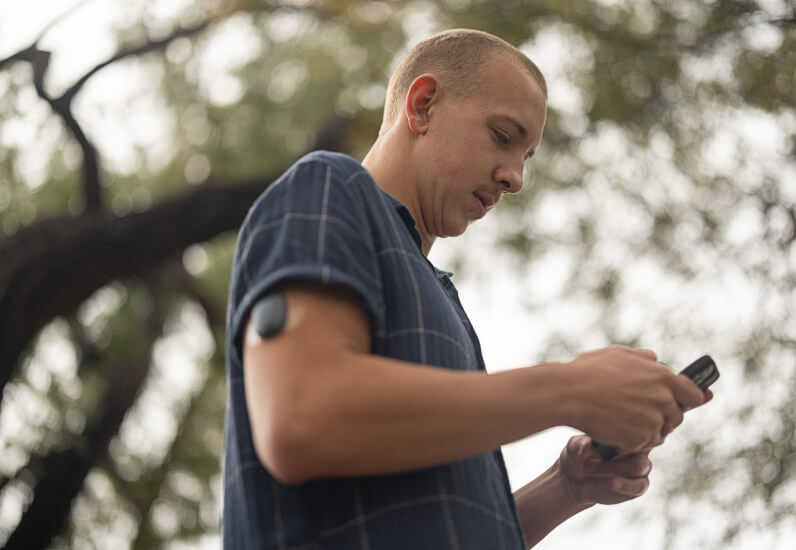 Man using smartphone outdoors, wearing a blue plaid shirt, standing near trees with blurred green foliage background.
